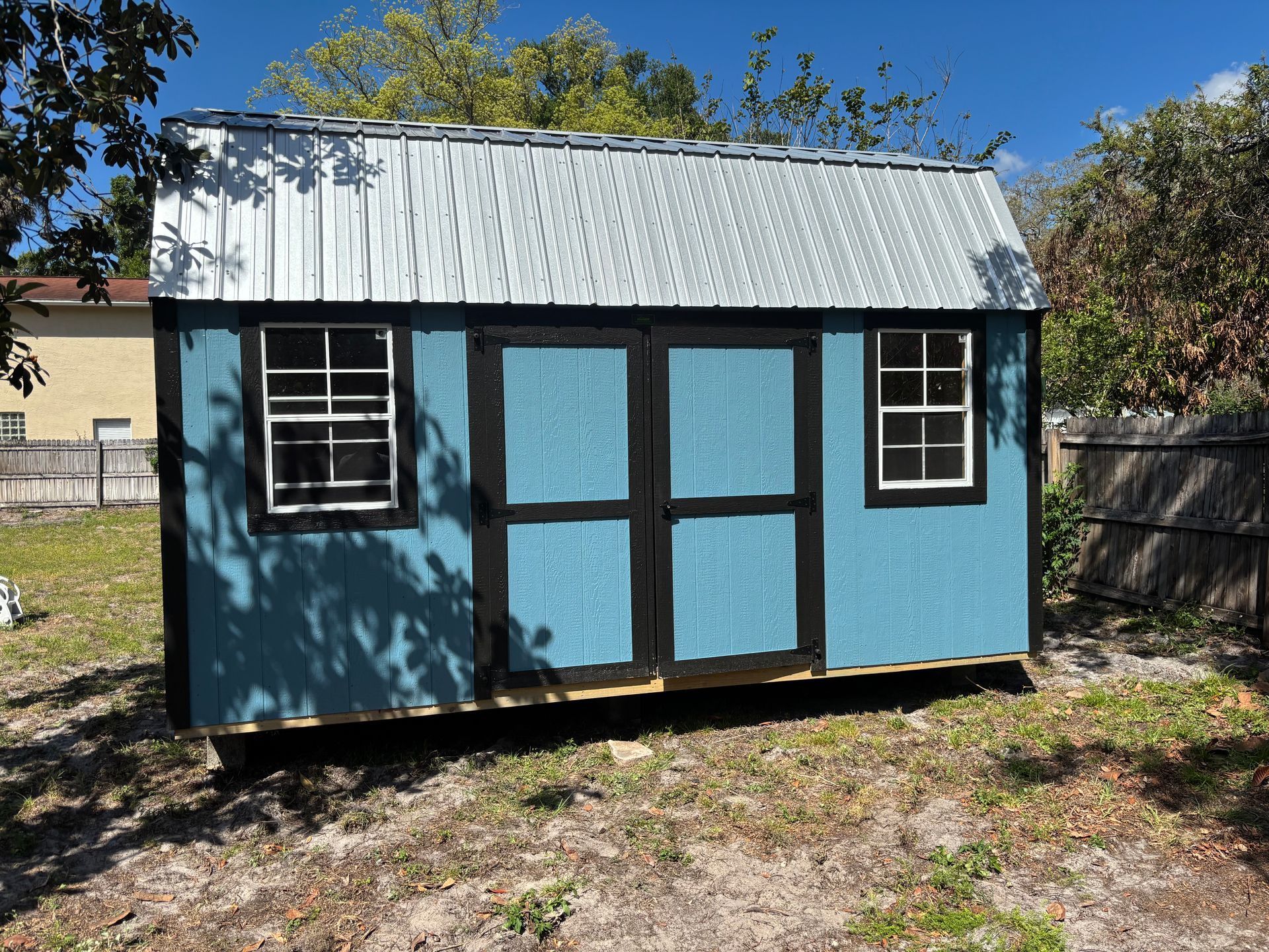 Small blue shed with a corrugated metal roof and white-trimmed windows in a sunny yard