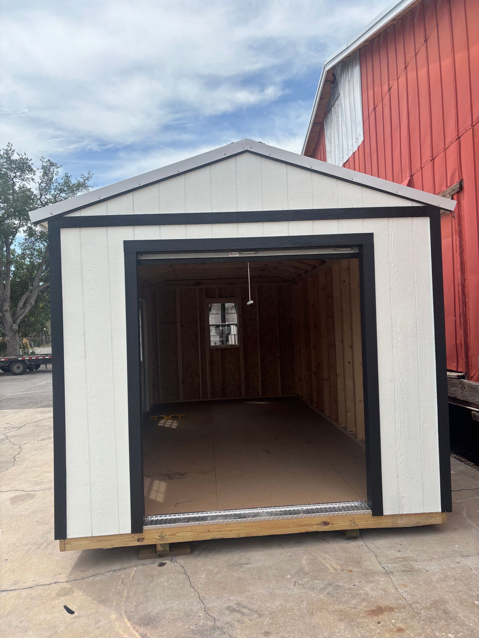 Open wooden shed with white and black trim, standing beside a red building outdoors.