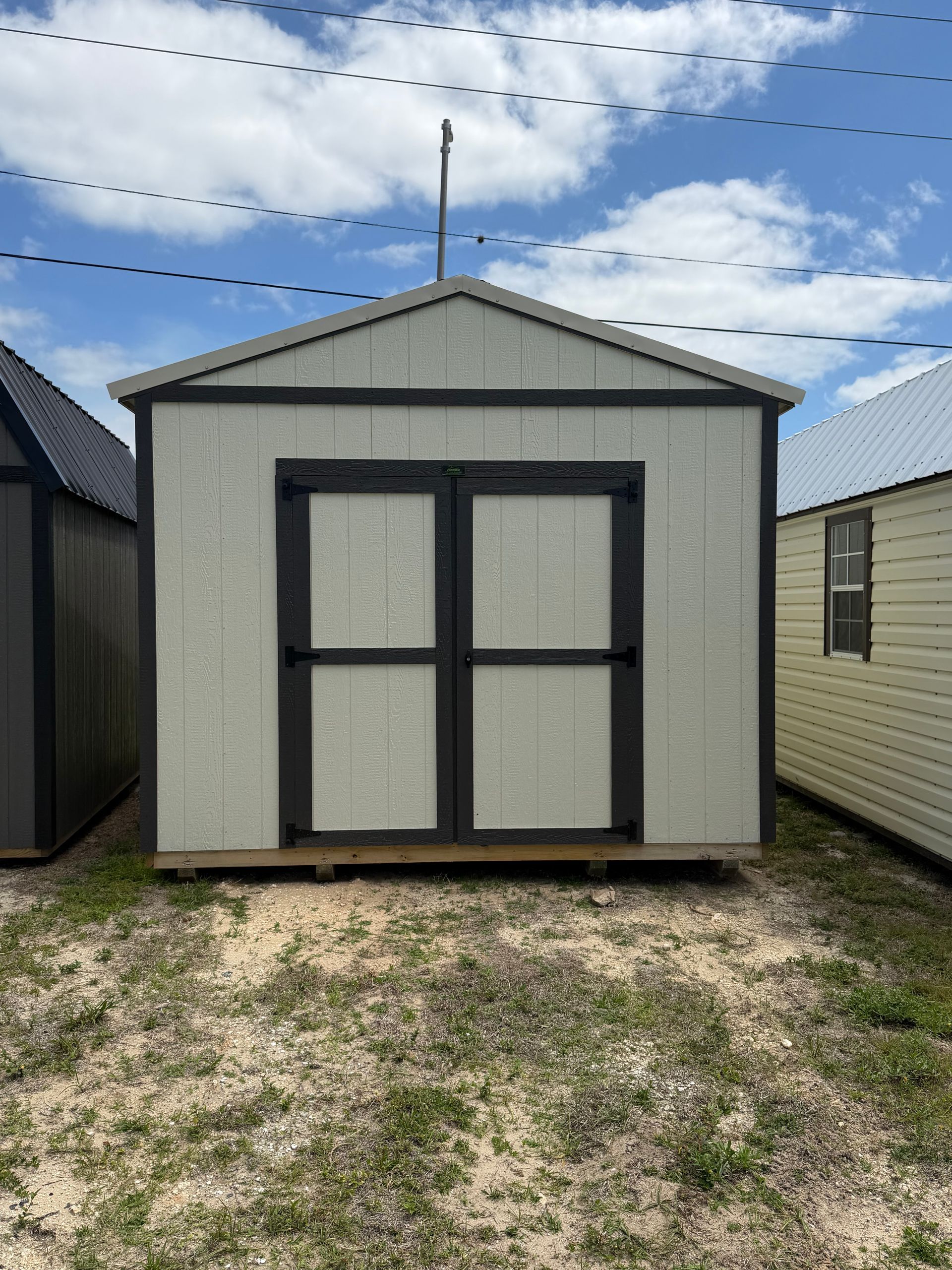 Small gray storage shed with black double doors on a grassy lot under a partly cloudy sky