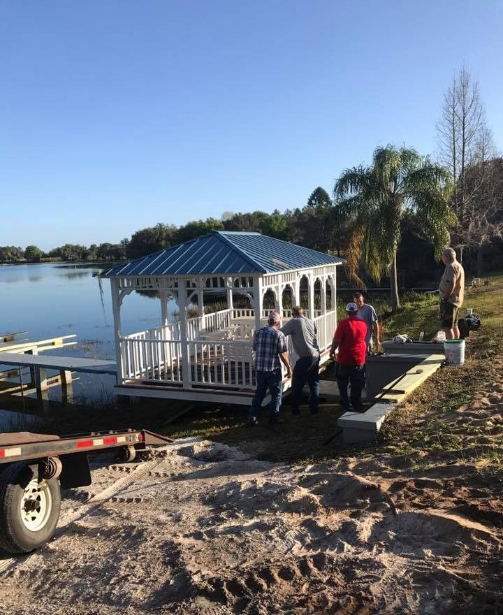 Workers move a white gazebo onto a lakeside dock beside calm water and palm trees.