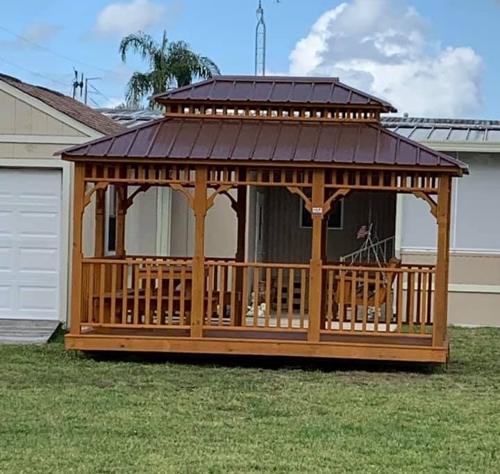 Wooden gazebo with brown metal roof and railings on a grassy lawn beside garages