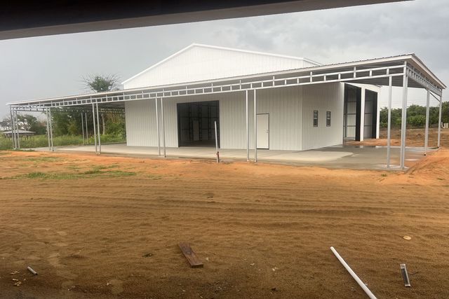 White building with a metal roof and open porch on a dirt lot, surrounded by sparse greenery