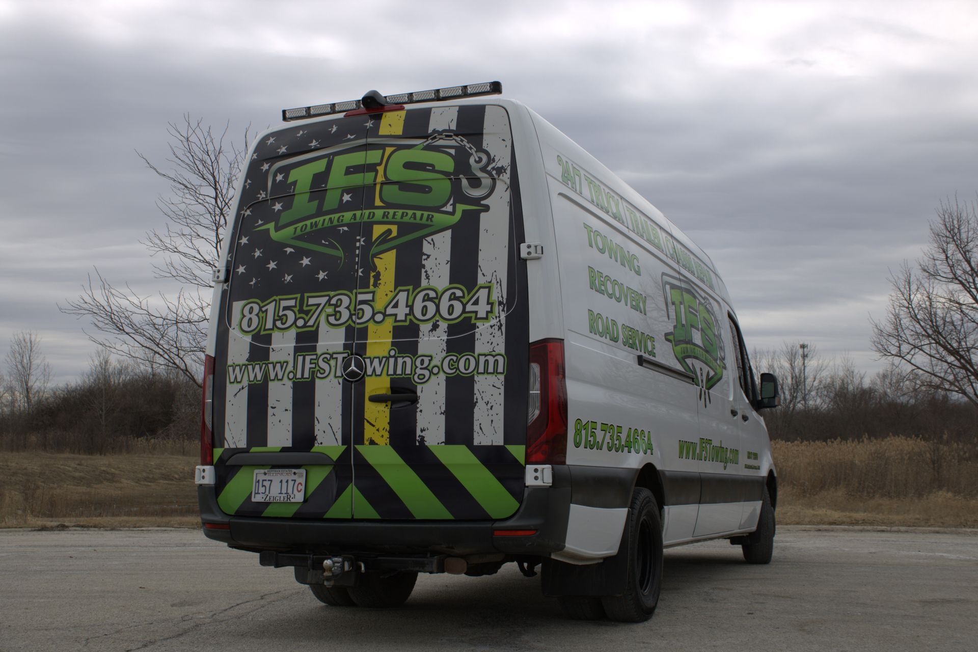 White van with IFS Towing logo and American flag design on the back. Parked outdoors under cloudy sky.