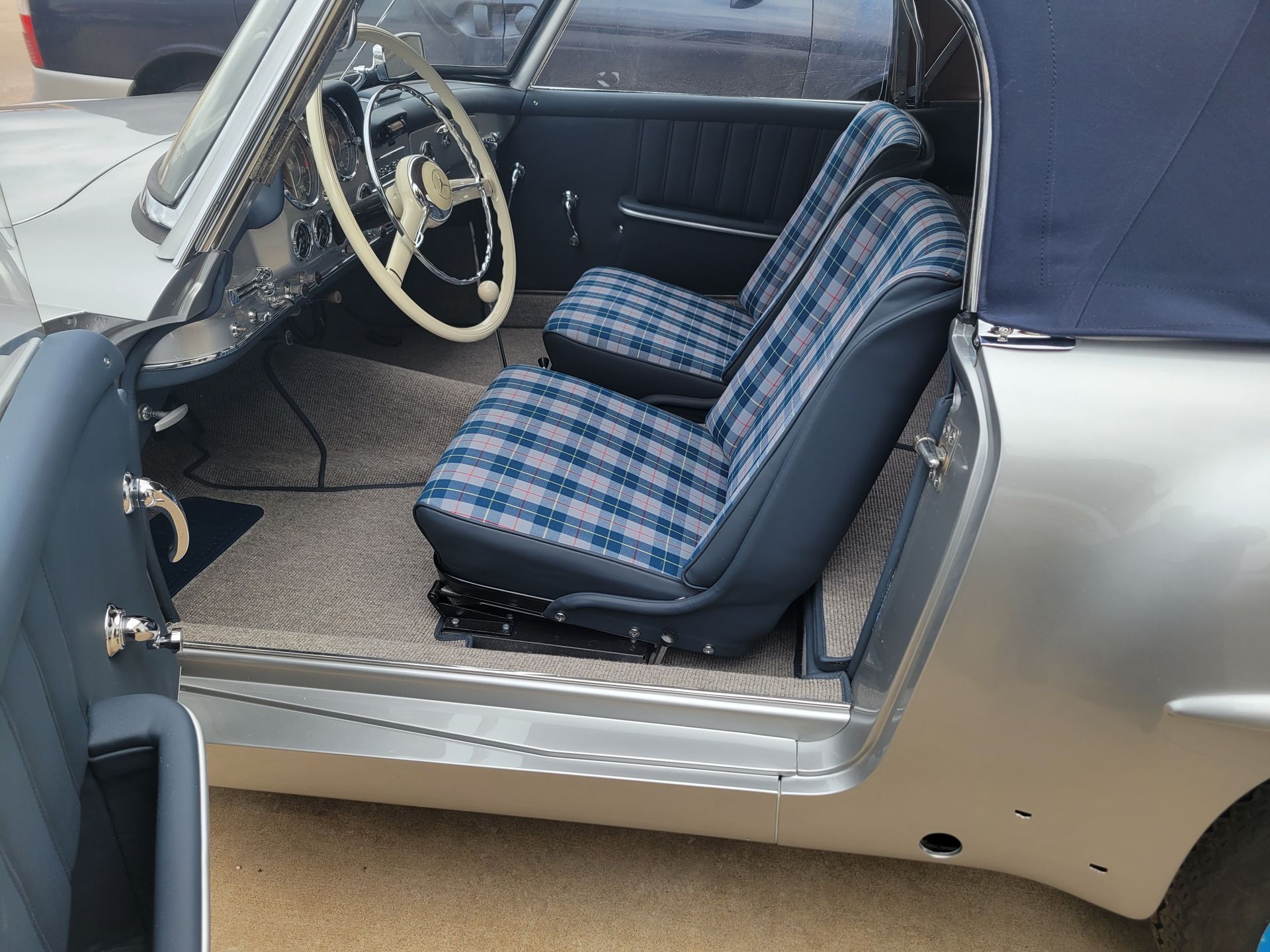 Vintage car interior with blue plaid seat upholstery and beige steering wheel, door open to the side