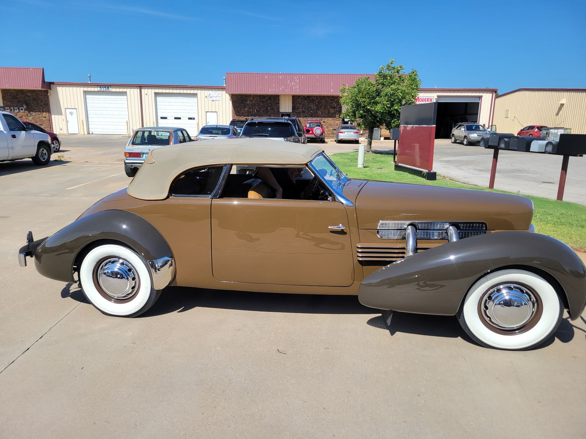 Brown vintage hot rod coupe with black fenders and whitewall tires parked outdoors in front of buildings