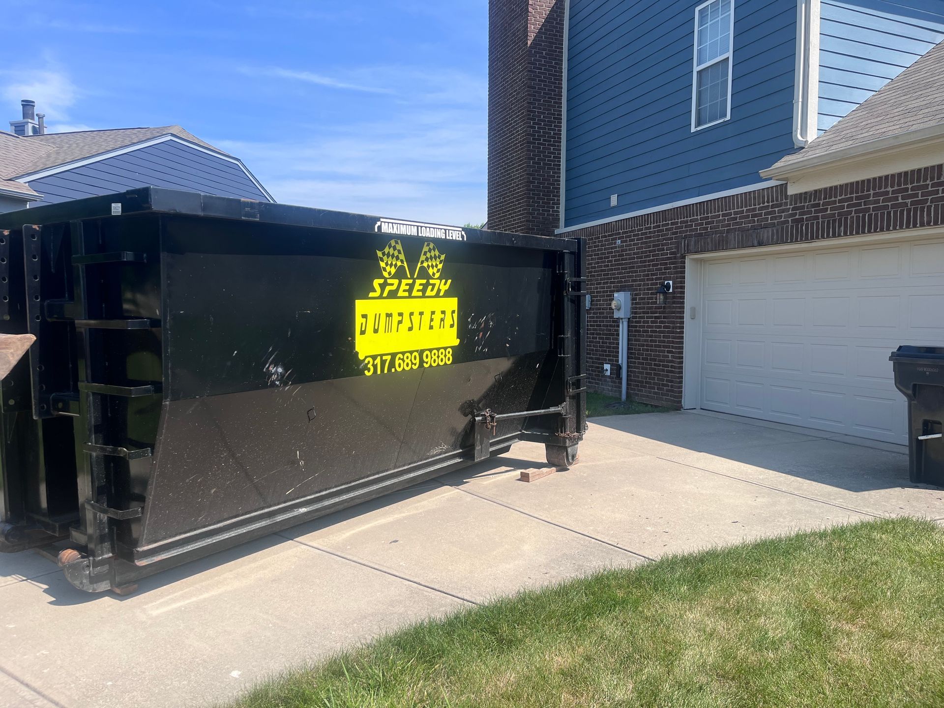 Black dumpster on a driveway in front of a blue house and garage on a sunny day.