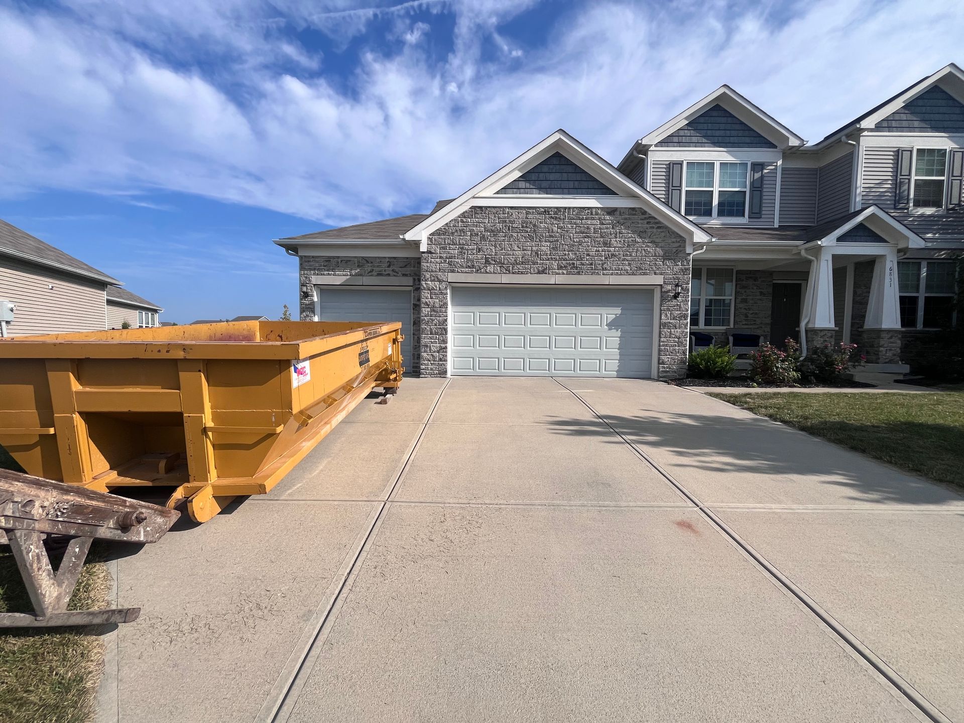 A large, yellow dumpster in front of a two-story house with a gray garage door and driveway on a sunny day.