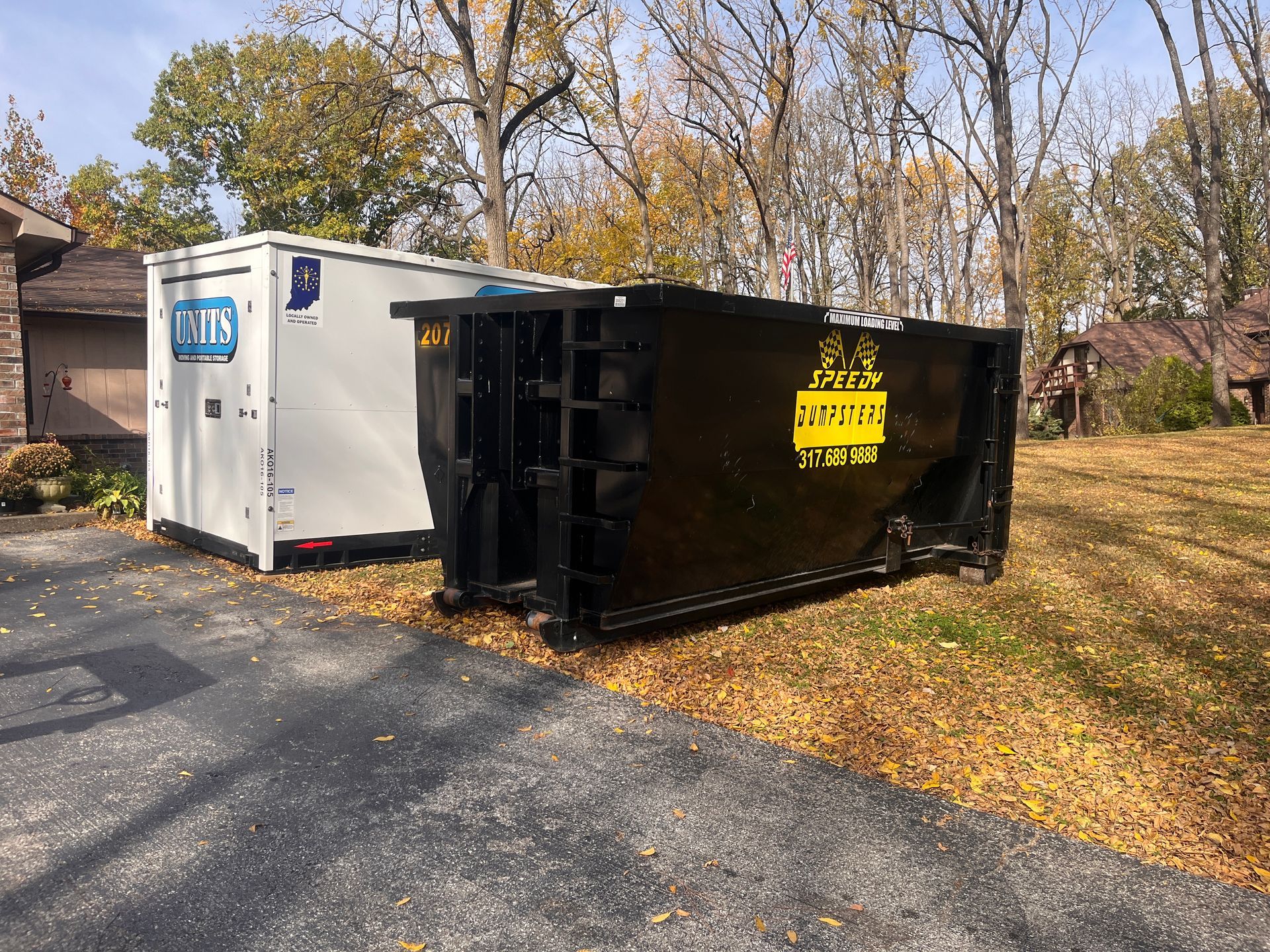 White truck and black dumpster on asphalt, with trees and a house in the background.