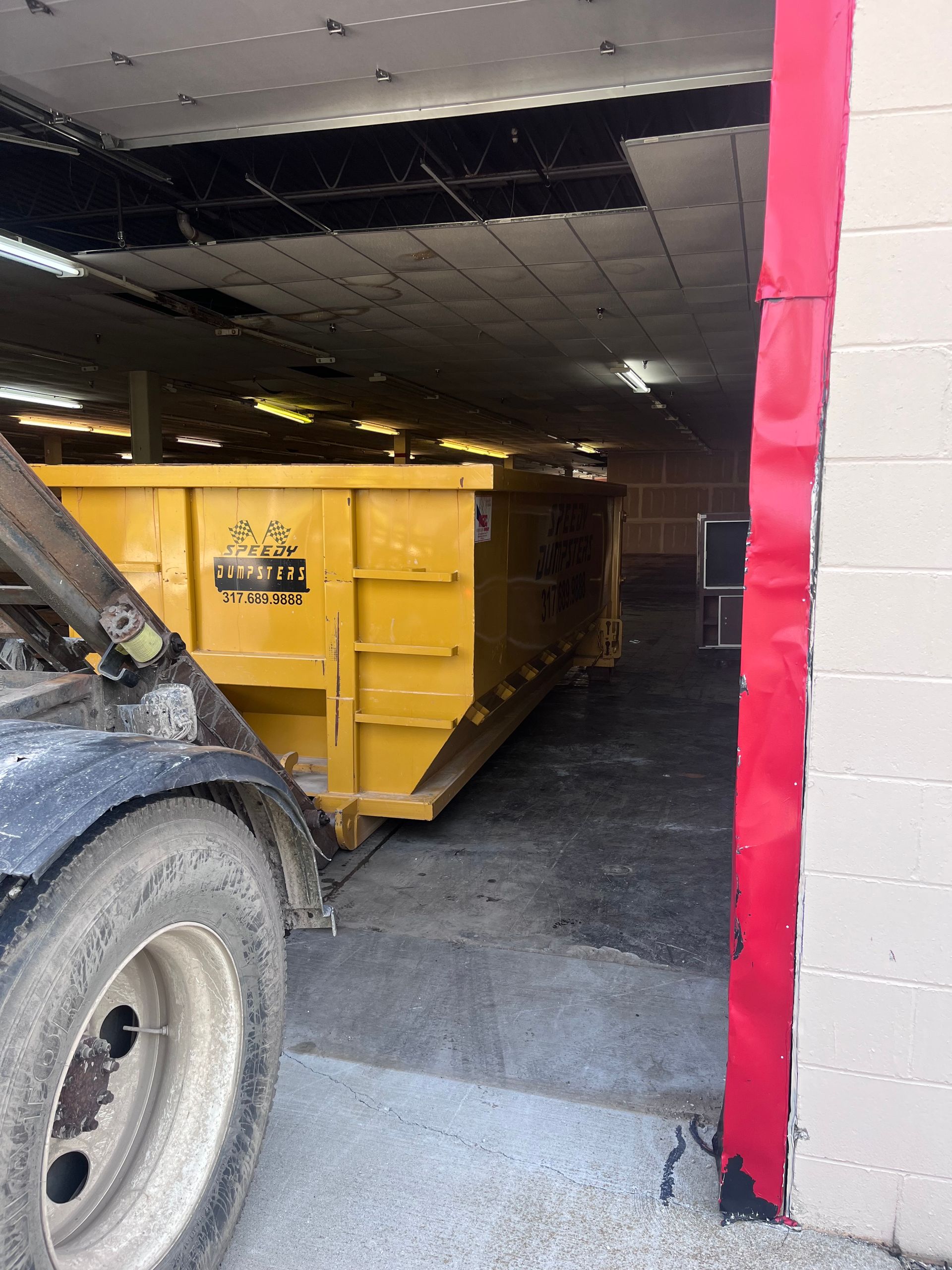 A yellow dumpster sits inside a building with a damaged ceiling. A truck is partially visible on the left.