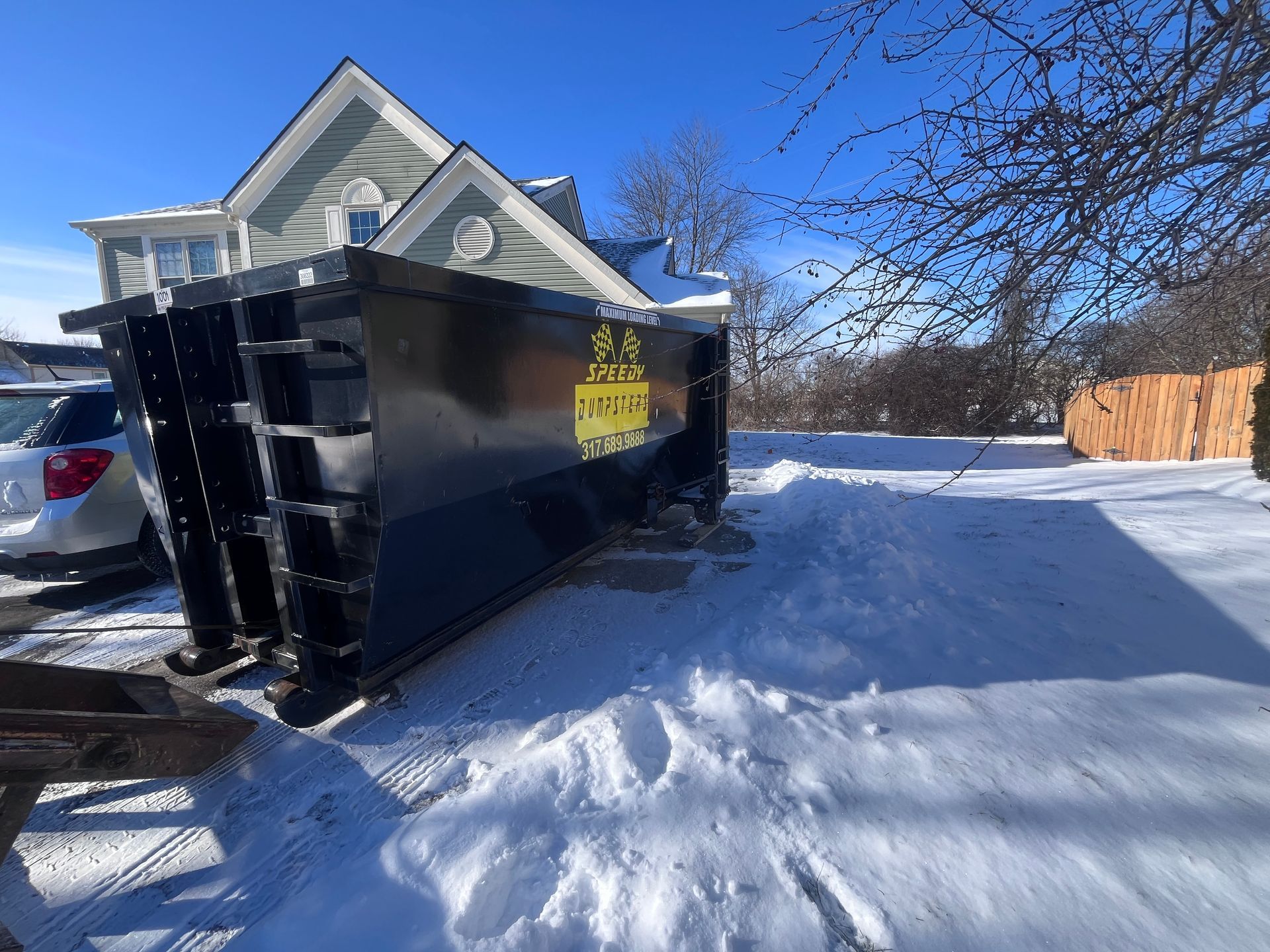 Black dumpster in snow-covered residential yard with a house and fence in the background.