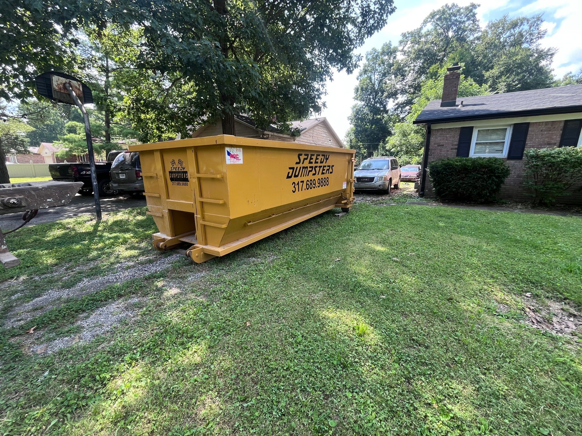 Yellow dumpster on a lawn in front of a house, parked next to a driveway and road.