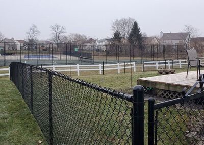 Black chain-link fence in foreground, tennis courts and white fence in background under overcast sky.