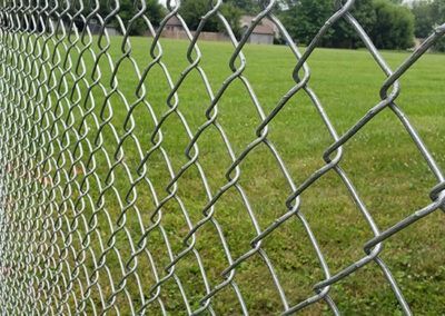 Chain-link fence in front of a green grassy field, trees, and buildings in the background.