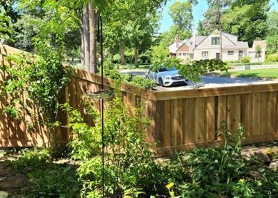Wooden fence in a sunny residential area. A car is parked on the street near a house in the background. Green foliage grows along the fence.