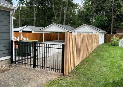 Black metal gate opens to a paved area with a green trash bin. Wooden fence to the right, white garage and carport in the distance.