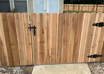 Wooden gate with black hardware and decorative top, set in a wood fence with a stone path.