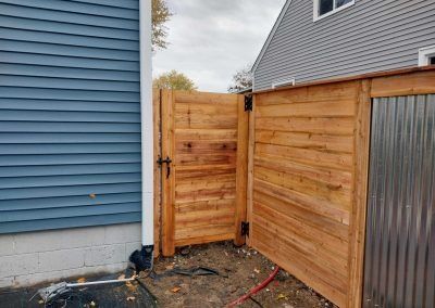 Wooden privacy fence with a gate next to a blue house and a corrugated metal structure.