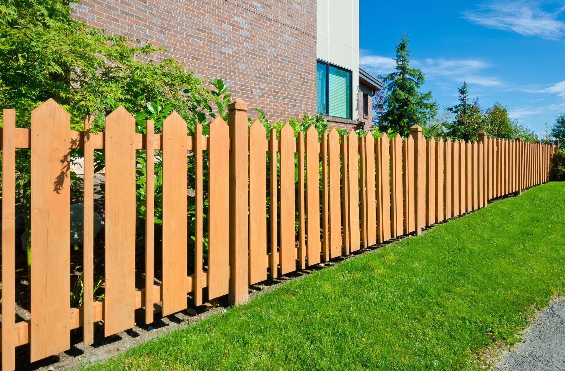 Wooden picket fence along a green lawn, with a brick building and blue sky in background.