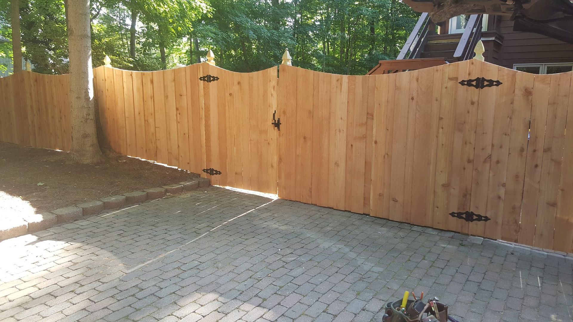 Wooden privacy fence along a paved backyard patio, with trees and sunlight in the background.