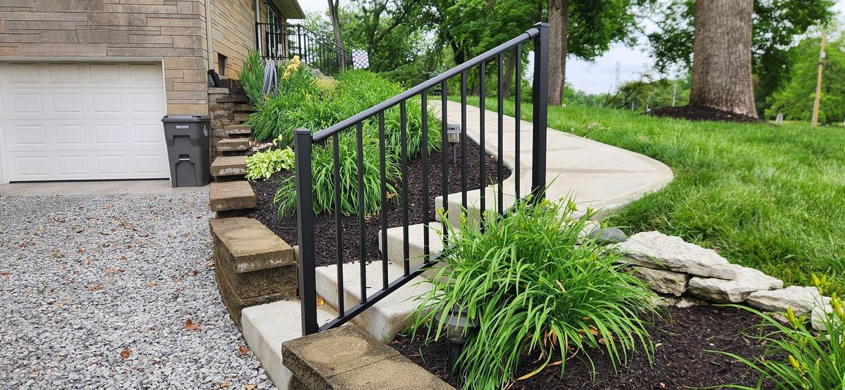 Front steps with black metal railing beside a stone house, leading to a curved walkway and landscaped garden.