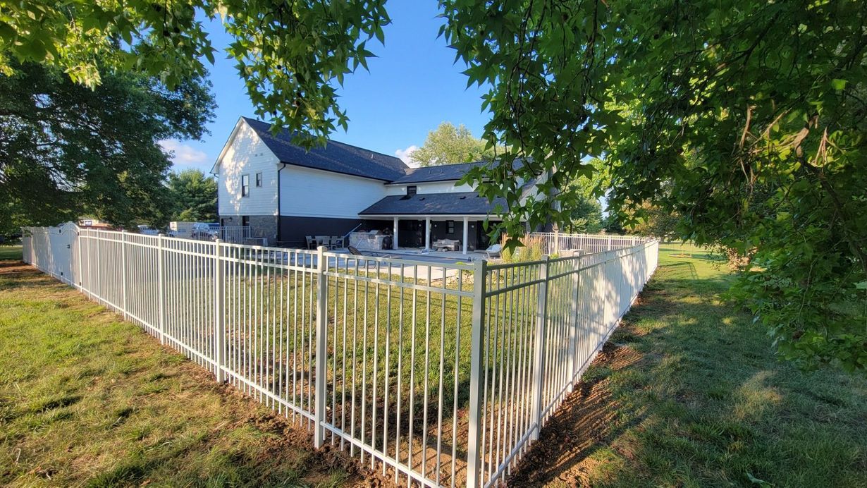 White fence around a grassy yard beside a house under leafy trees on a sunny day