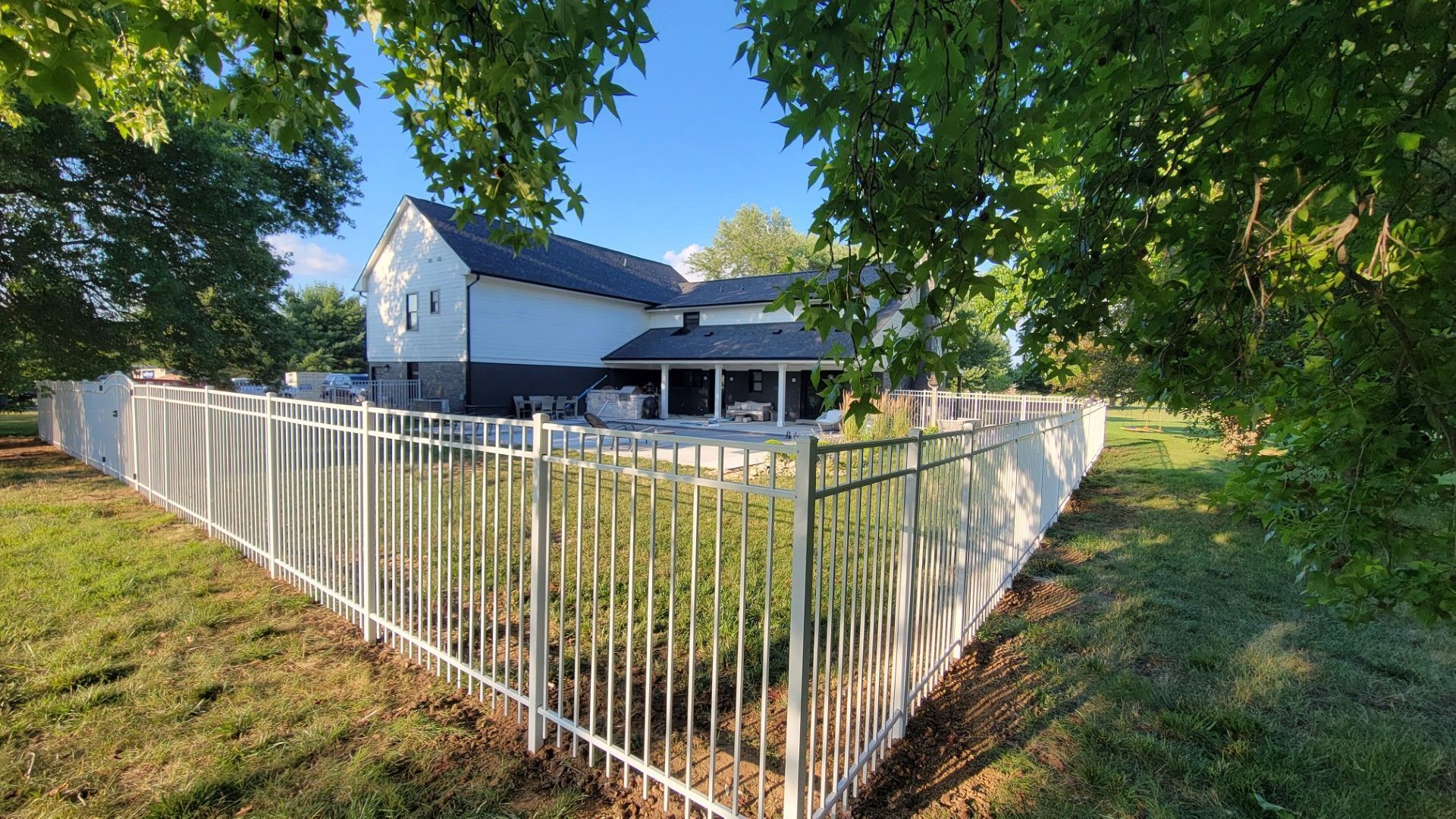 White fence around a grassy yard beside a house under leafy trees on a sunny day