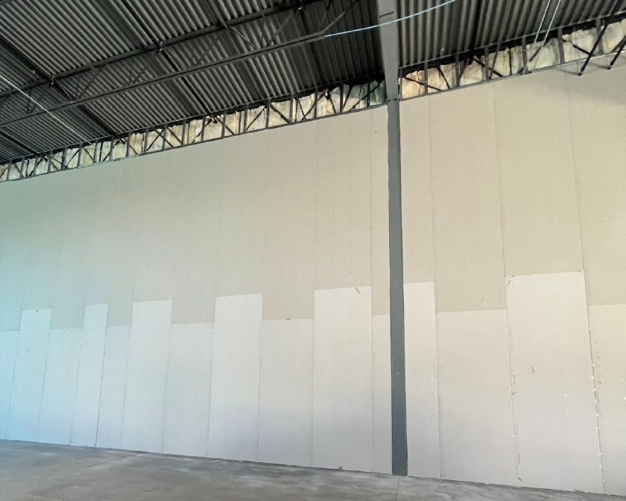 Interior of a large industrial building with light-colored drywall covering a wall; corrugated metal roof visible.