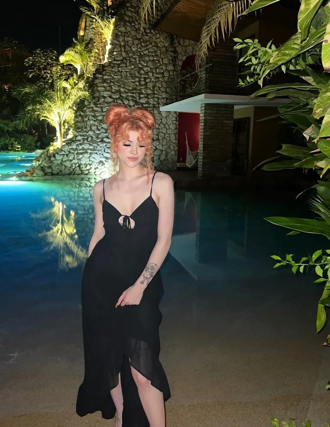 a woman in a black dress is standing next to a pool at night