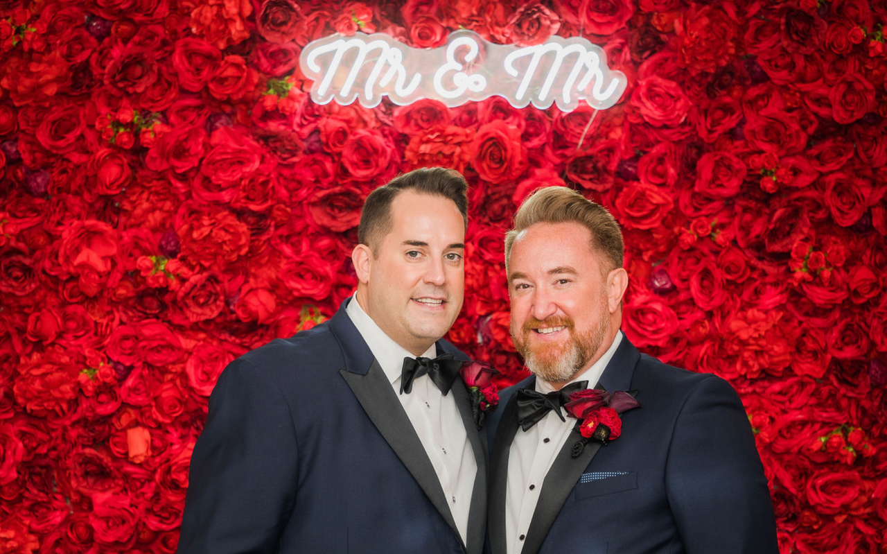 two men in tuxedos are posing for a picture in front of a wall of red roses