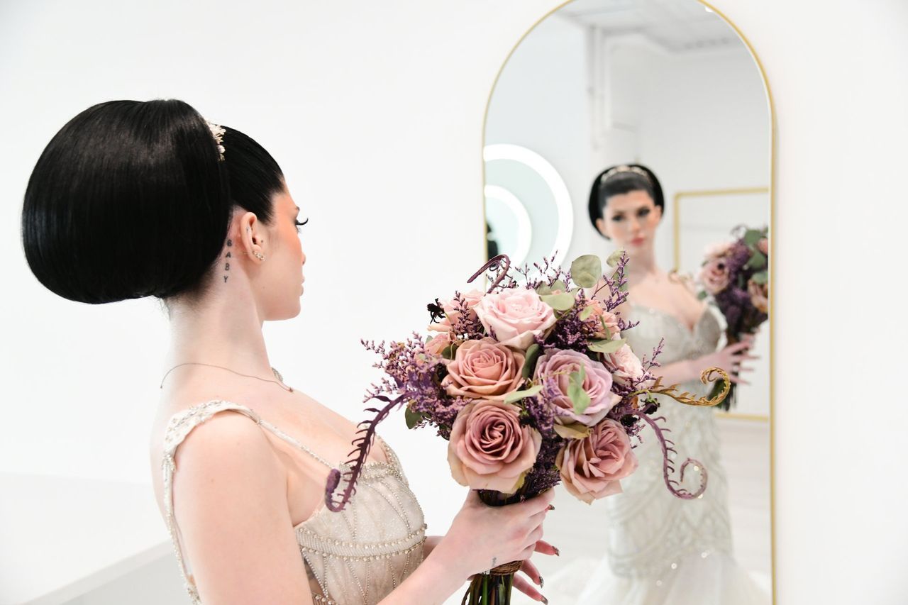 a woman in a wedding dress is holding a bouquet of flowers and looking at herself in the mirror