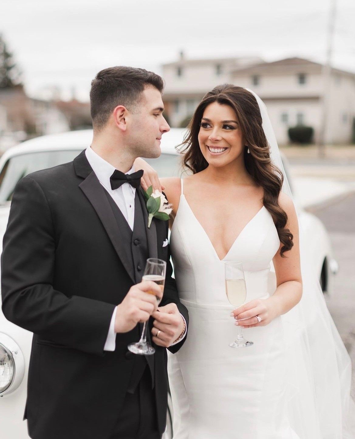 a bride and groom are standing in front of a white car holding champagne glasses
