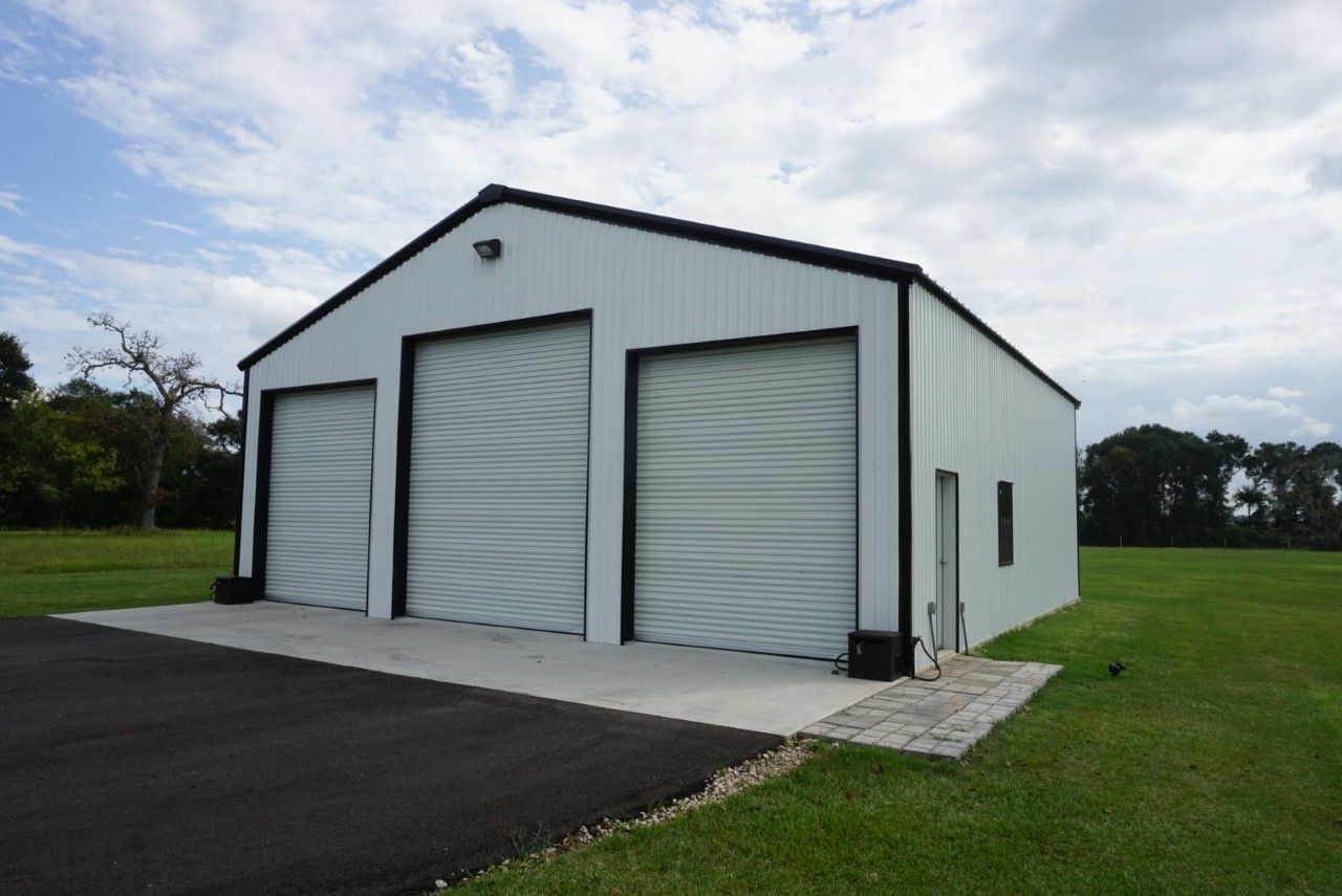 A white garage with three garage doors is sitting in the middle of a grassy field.
