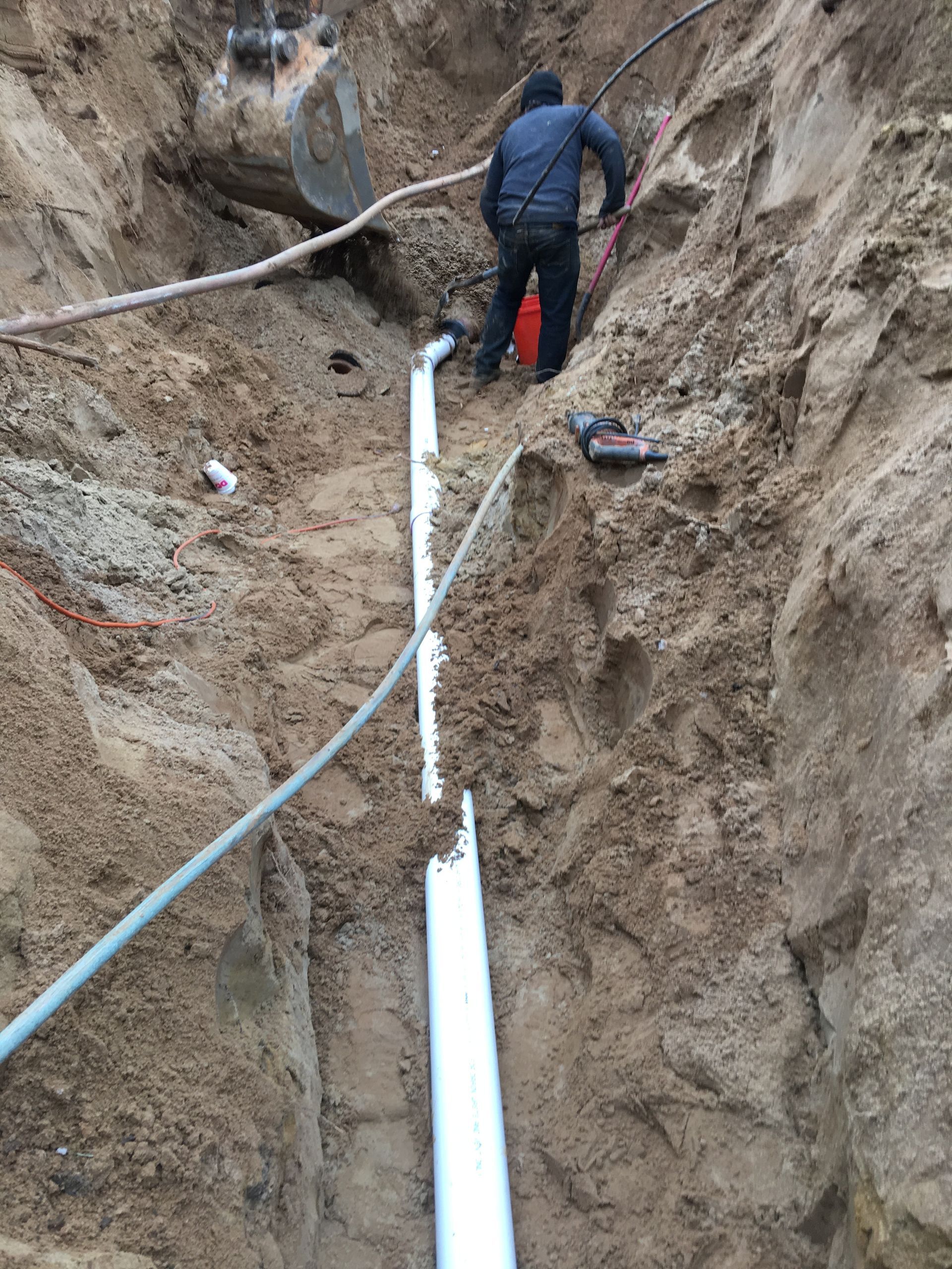 Worker in a trench installing white pipes along a dirt excavation site.