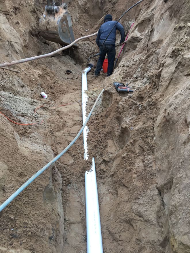 Worker in a trench installing white pipes along a dirt excavation site.