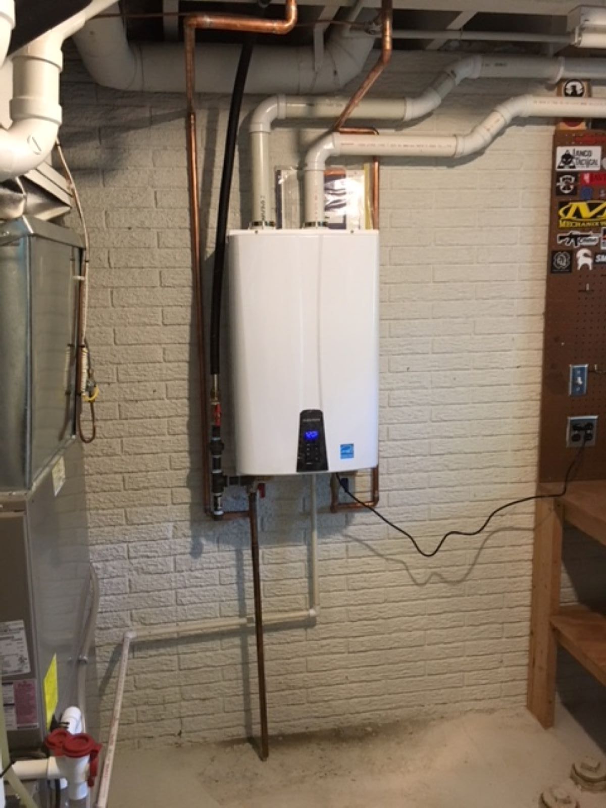 Wall-mounted white water heater in a basement utility room with exposed pipes and brick wall.