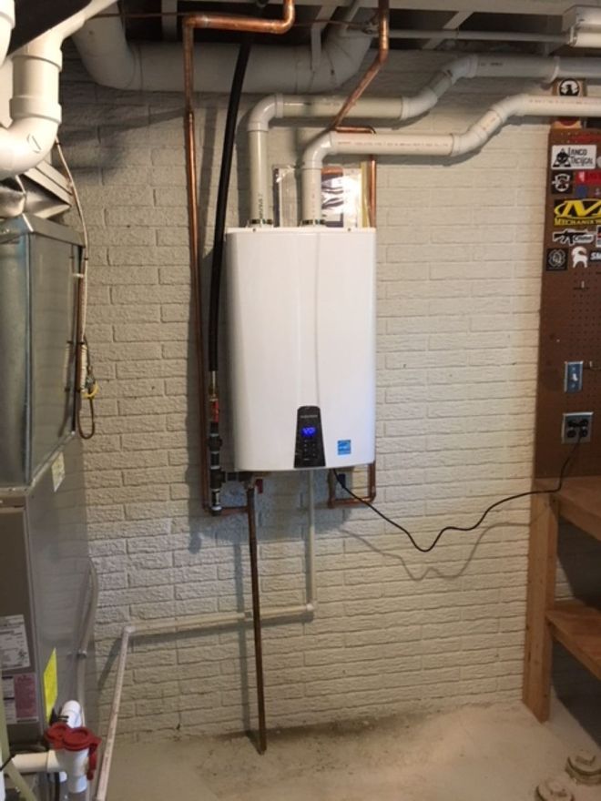 Wall-mounted white water heater in a basement utility room with exposed pipes and brick wall.
