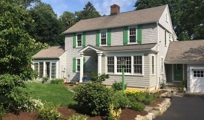 A light-gray two-story house with green shutters, a central portico entrance, and a garage wing set in a lush garden.