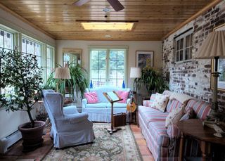 Cozy sunroom with floral sofa, blue chair, indoor plants, and large windows under a wood ceiling