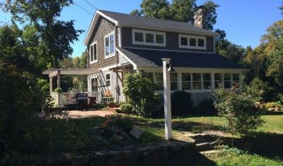 A two-story gray house with a porch, chimney, and a stone retaining wall in a sunlit, wooded yard.