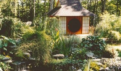 A small Japanese-style tea house with a circular door, nestled in a lush, green woodland garden with a foreground pond.