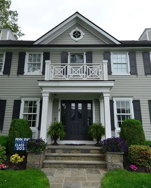 Gray house with black shutters, balcony, and front door; stone steps lead up to the entrance.
