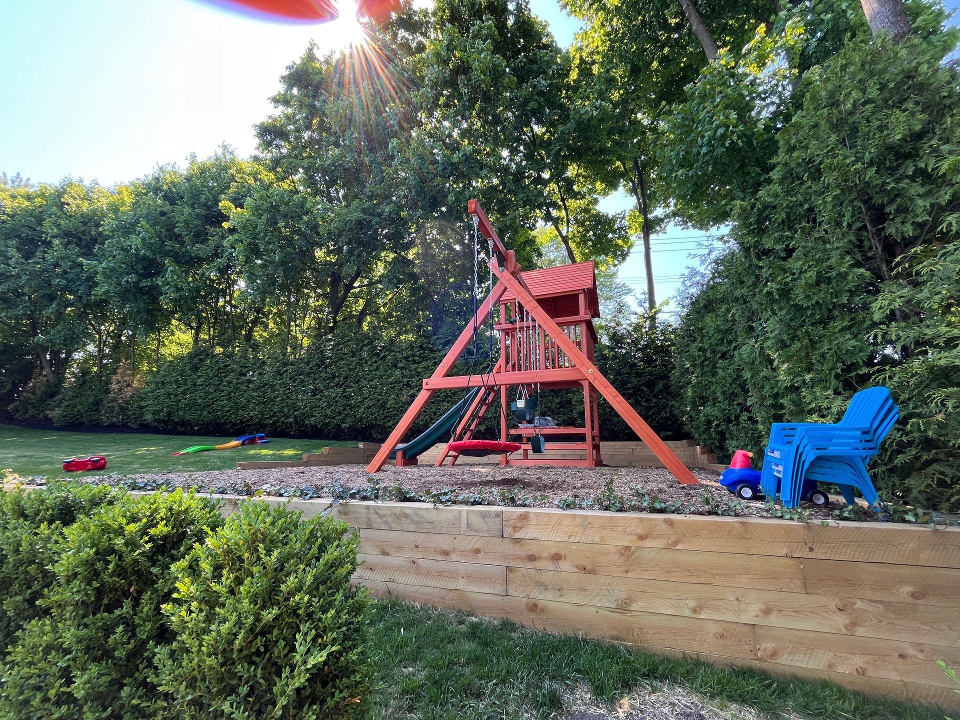 Backyard with a red A-frame play structure and blue chair beside a raised garden bed and trees