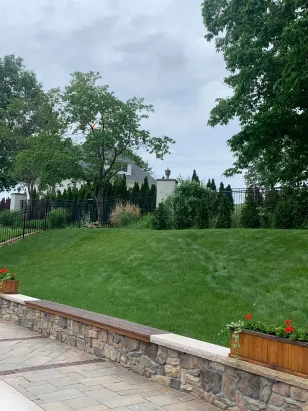 Lush green lawn with stone wall and flower boxes. Trees and a house in the background under a cloudy sky.
