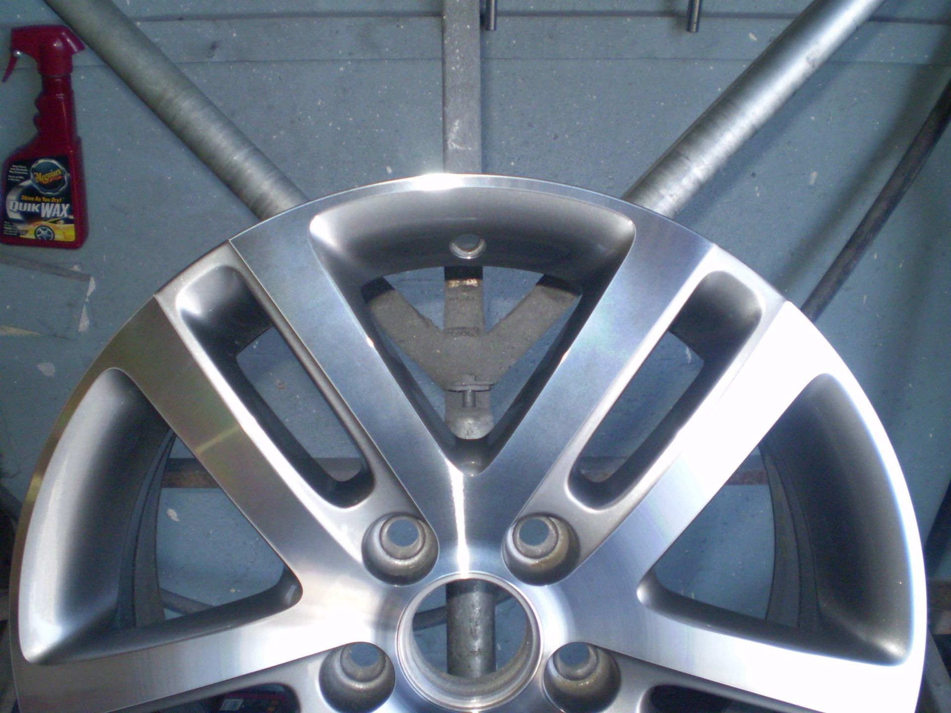 Silver car wheel suspended in front of a metal frame, with a cleaning product visible on the wall.
