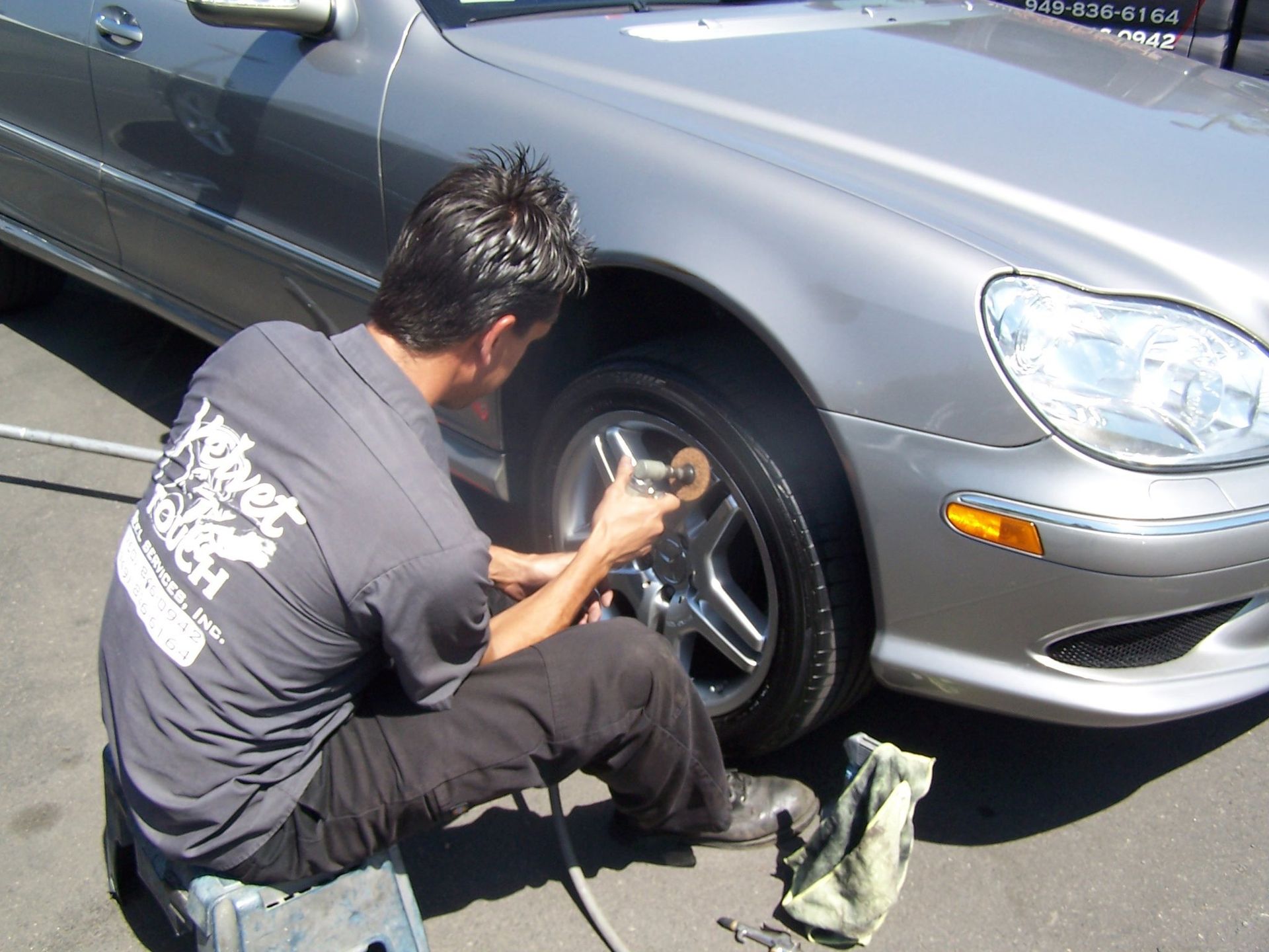 Man cleaning the chrome wheel of a silver car outside, using a power tool and cloth.