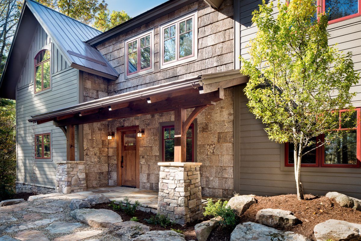 A large house with a stone facade and a metal roof is surrounded by trees and rocks.