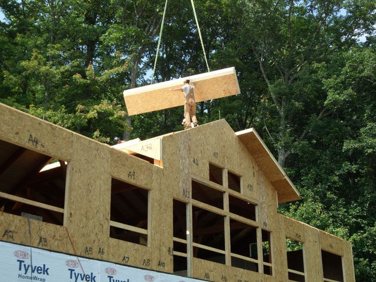 A crane is lifting a piece of plywood on top of a house under construction.