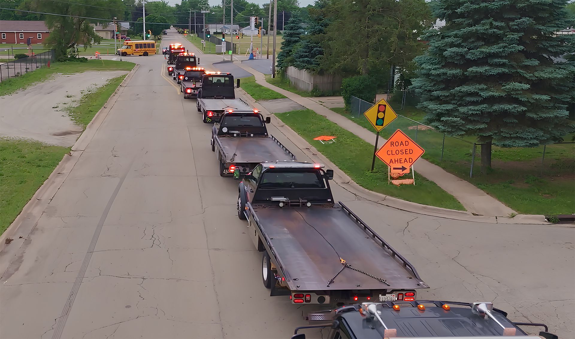 A long line of flatbed tow trucks, some with flashing lights, driving down a street. A school bus is in the distance.