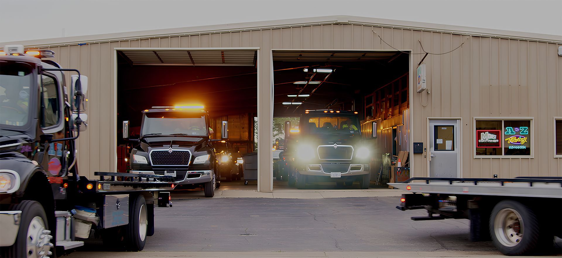 Tow trucks parked outside a garage. Headlights shine.