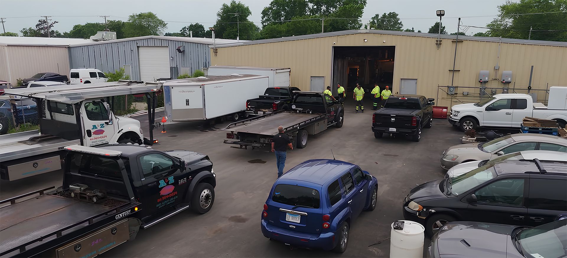 A tow yard with several flatbed tow trucks, a white trailer, and a blue SUV. A group of people are by a large open door.